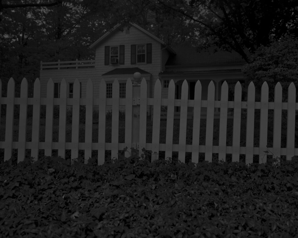 Dark black and white photograph of white picket fence with house behind